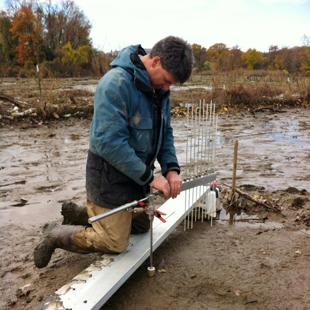 Field technician uses equipment to monitor marsh elevation while kneeling in a muddy bank.