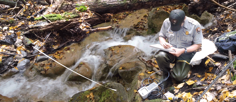 A NETN water quality tech kneels in a rushing stream while writing on a datasheet.