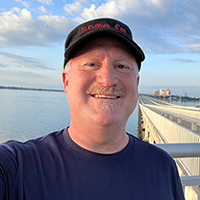 Eliot Rendleman, NPS Writer-Editor, wearing a black hat, standing on a bridge over Biloxi Bay, smiling at the camera. The background shows the coastline of Biloxi, Mississippi.