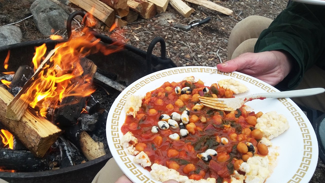 Camp meal A hand holding a white plate with food and a fork on it and a campfire in the background.