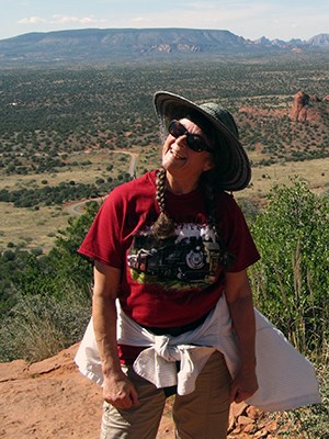 Woman with hat,red shirt, smiling with mesa in the distance and valley below.