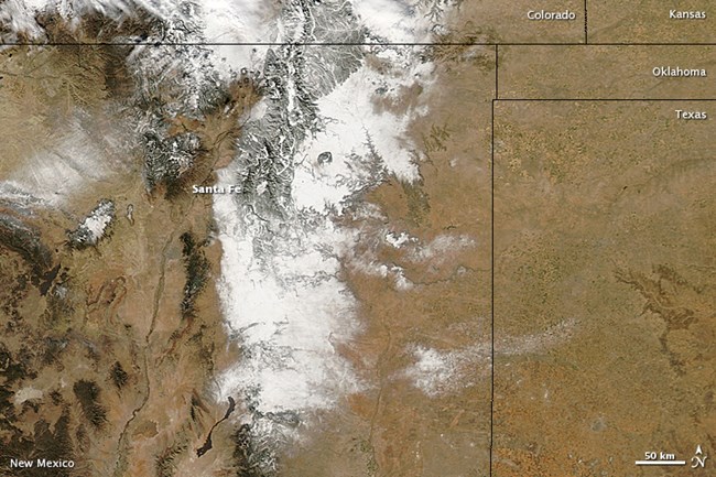 MODIS_NewMexicoSnow_Phenology Satellite image of New Mexico and surrounding states. The background is mostly brown with dark green areas that represent forests. White areas cover the central area of the image.