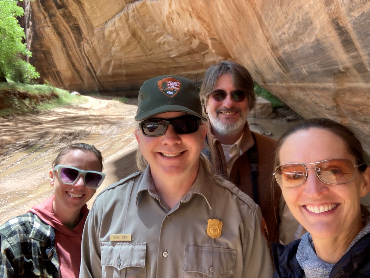 MegWhiteVisit_CACH_2024_4 Four people grouped together and smiling in a dry wash at the bottom of a sandstone canyon.