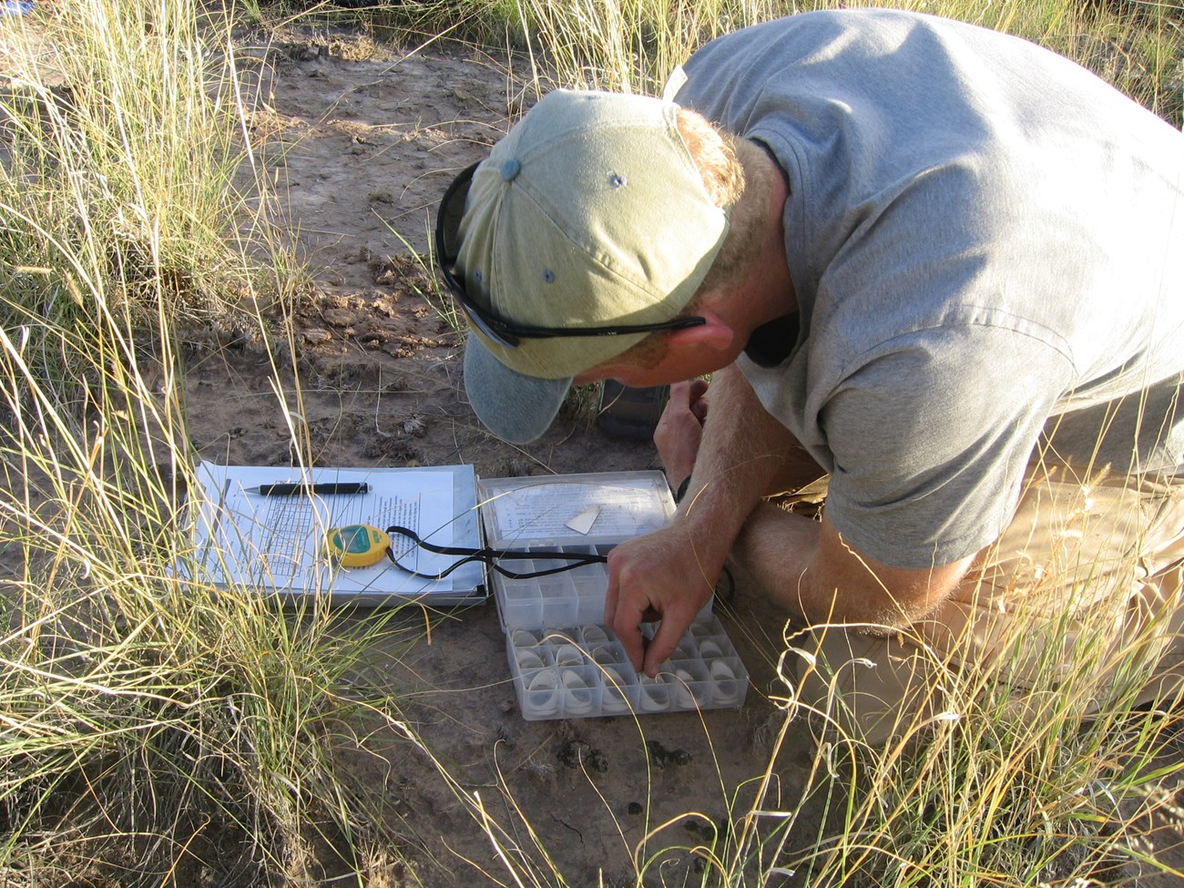 A person conductin soil stability tests with a plastic box that holds the soil, a yellow stopwatch, and a clipboard with paper on it.