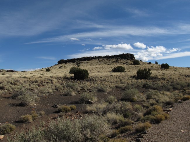 Expansive grass and shrubland in front of ancestral Puebloan structure beneath a partly cloudy blue sky