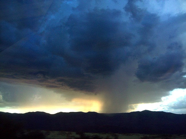 Dark billowy clouds with localized rain over a mountainous landscape.