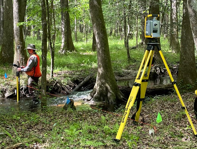 Tripod in foreground, man in uniform holding pole in a stream to the left