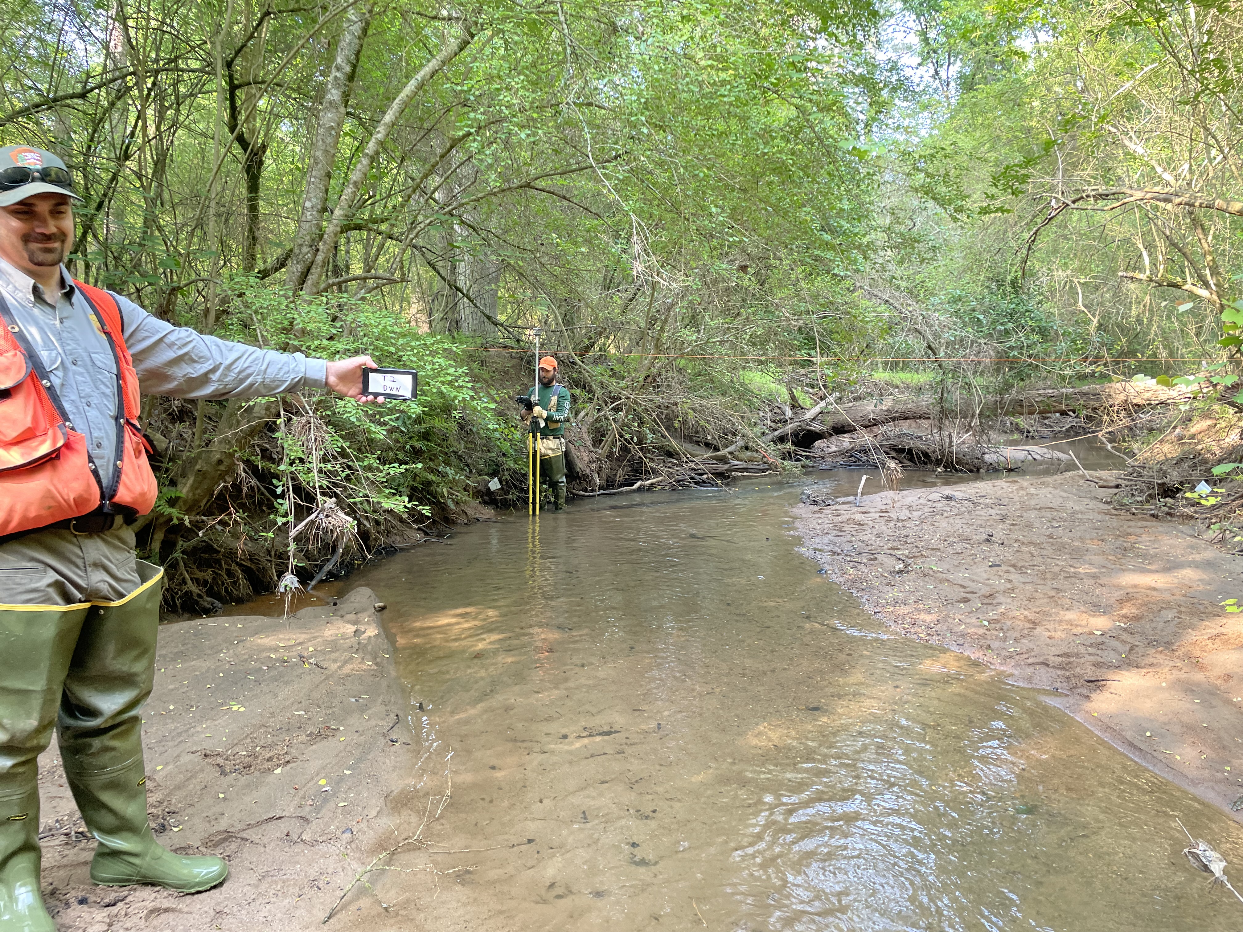 Man in hip waders standing next to stream holding a placard with writing
