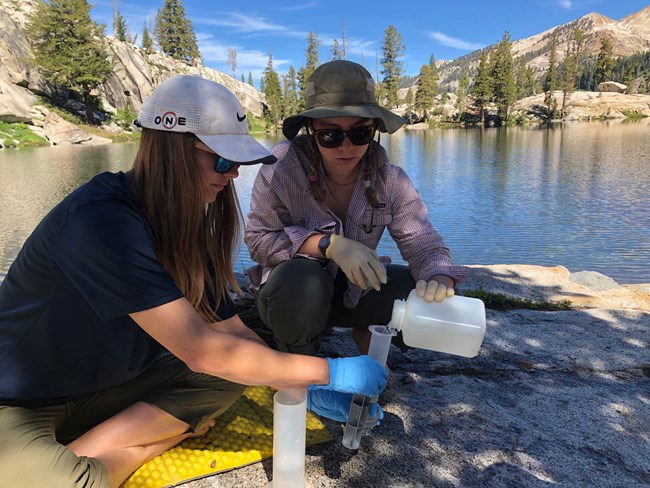 Two women sit at the edge of a lake - one pours a lake water sample from a plastic bottle into a filter held by the other woman.
