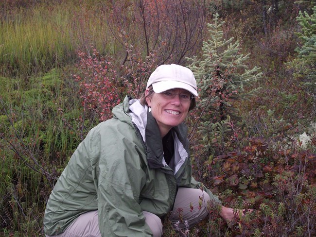 Woman poses smiling in front of tundra shrubs and small tree.