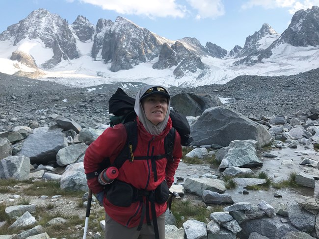 Erika Blomdahl, Ecologist Woman wearing hat and warm jacket stands wearing a pack with a view of rocky slopes and Wind River Mountains in background.