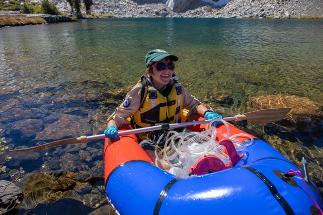 Lake monitoring crew member prepares to collect mid-lake sample. Woman sits in raft with monitoring equipment, preparing to paddle out to middle of lake where she will collect water samples.