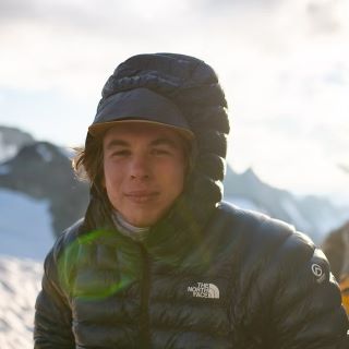 Ben, a mid-twenties man with mid-length blonde hair, wearing a dark, puffy winter coat with the hood up in front of rugged mountains covered in snow. Ben, a mid-twenties man with mid-length blonde hair, wearing a dark, puffy winter coat with the hood up in front of rugged mountains covered in snow.
