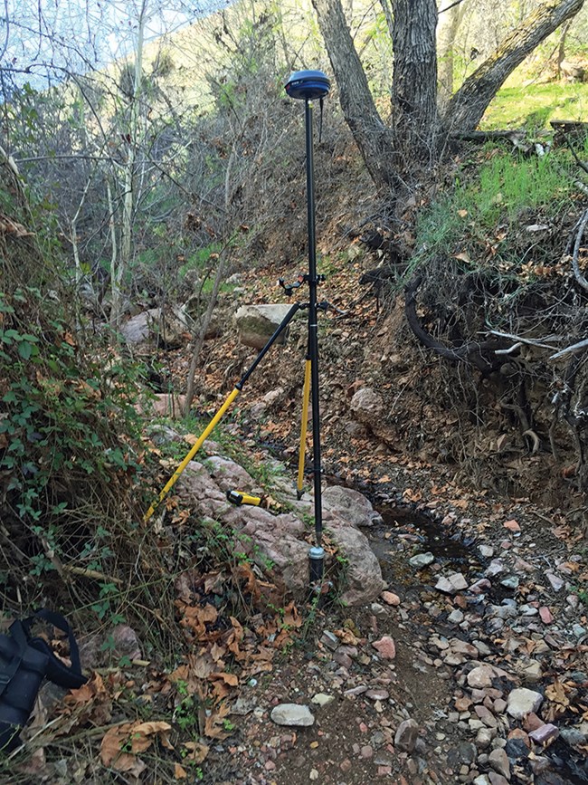 Pod-shaped apparatus sits atop a pole next to a monitoring well near a small riparian channel.