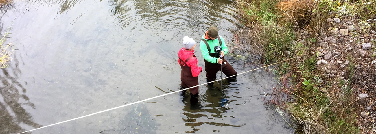 Flow monitoring Camera looks down on two people in neoprene waders measuring streamflow next to a transect tape