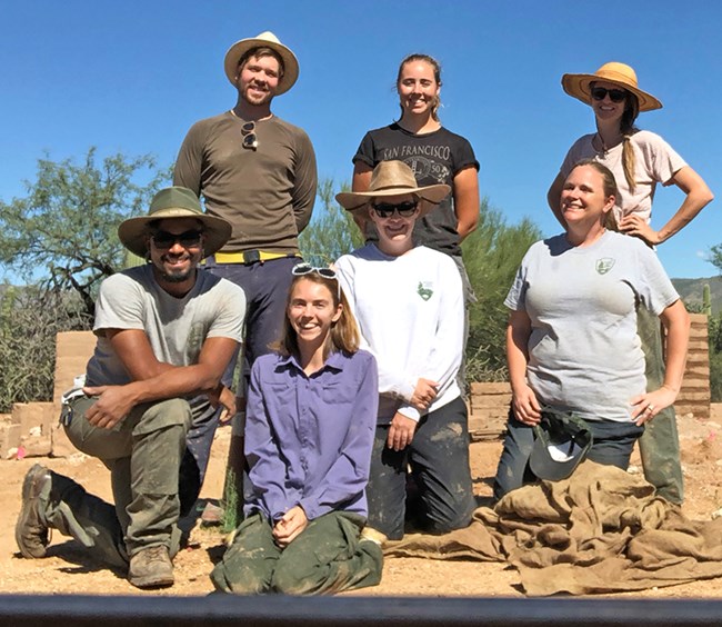 A group of people in work clothes stand against a blue sky, smiling at the camera
