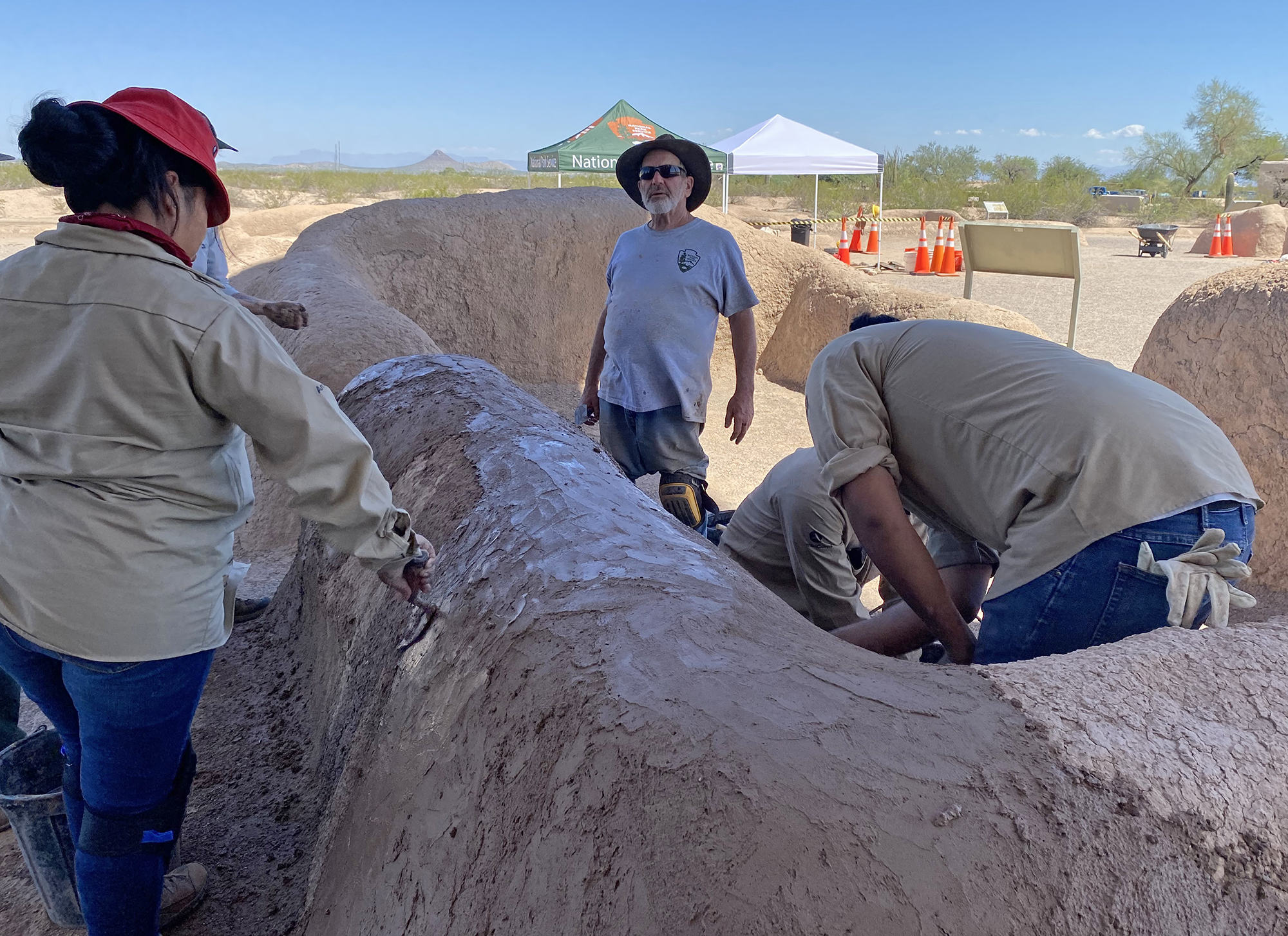 A woman uses a trowel on an adobe wall whose top and sides are partially covered in a new treatment.