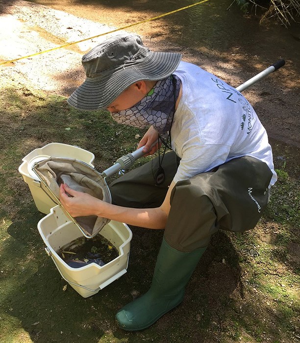 Woman wearing a gaiter over nose and mouth crouches in a stream