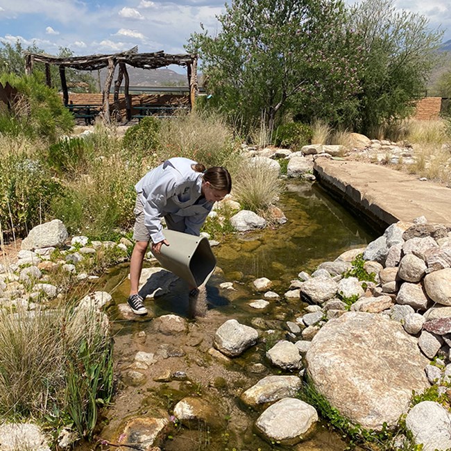 A woman pours sand from a bucket into an artificial stream