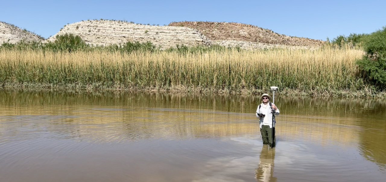 Man stands in a stream flowing through a meadow. He holds a metal rod with a disc on top.