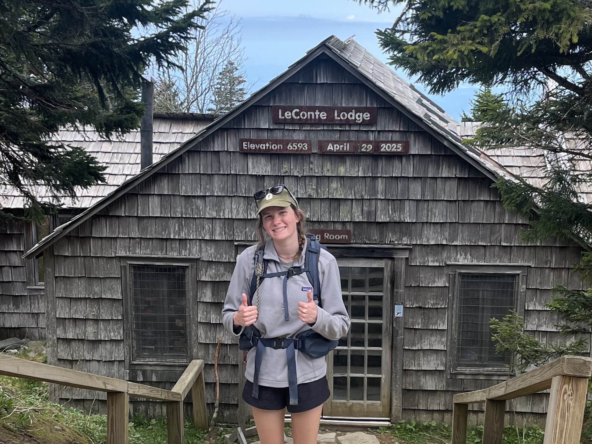 Kaitlin, a young woman smiling and giving two thumbs up. She's wearing a large backpack and standing in front of a wood cabin. Kaitlin, a young woman smiling and giving two thumbs up. She's wearing a large backpack and standing in front of a wood cabin.