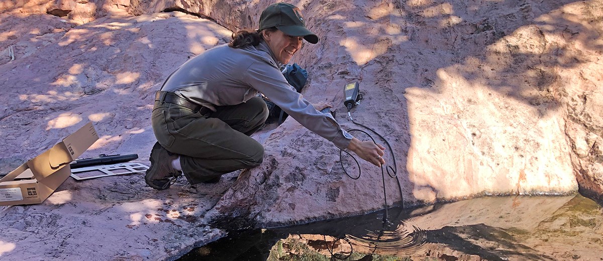 Woman in NPS uniform crouches next to tinaja, holding a probe wired to an electronic unit.