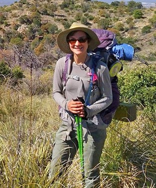 A woman wearing a backpack, hat, sunglasses, and hiking clothes stands on a verdant desert hillside.