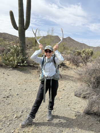 Savannah, a young woman standing in the desert near leafless shrubs and cactus, including a tall saguaro,smiling and holding a sun-bleached mule deer rack to her head. Savannah, a young woman standing in the desert near leafless shrubs and cactus, including a tall saguaro,smiling and holding a sun-bleached mule deer rack to her head.