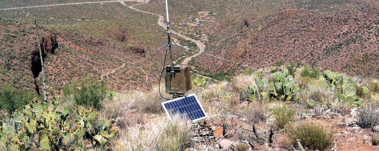 A metal box attached to a pole sits on a hillside high above the valley below. A solar panel powers the box.
