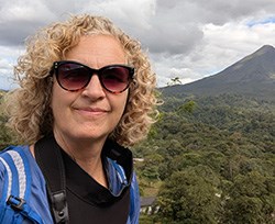 Tani Hubbard A woman with curly blond hair and sunglasses in front of a volcano surrounded by green trees.