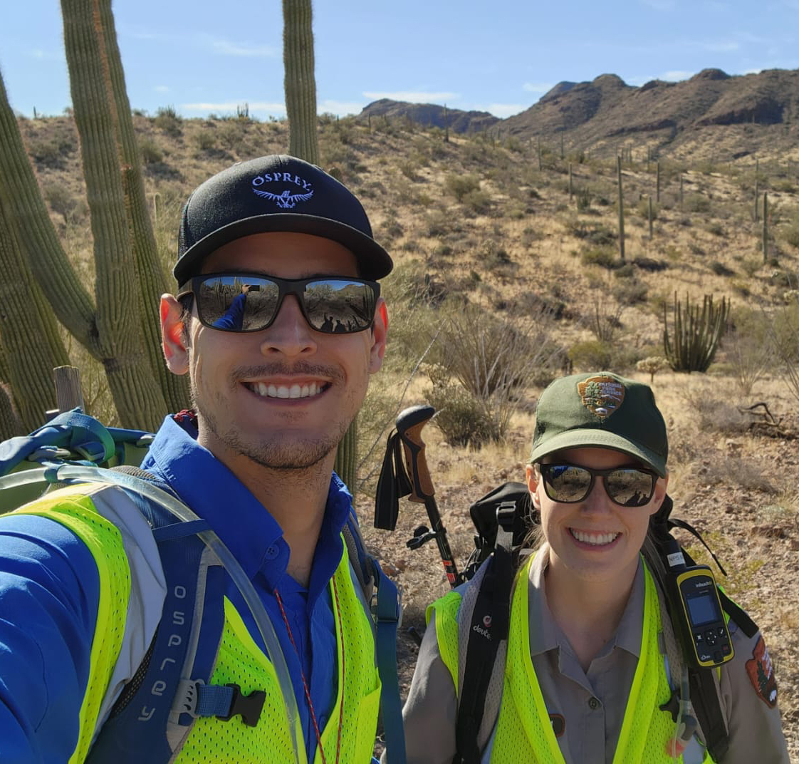 Smiling man and woman in sunglasses and yellow vests, organ pipe cactus behind and mountains in distance. Woman is in NPS uniform. Smiling man and woman in sunglasses and yellow vests, organ pipe cactus behind and mountains in distance. Woman is in NPS uniform.