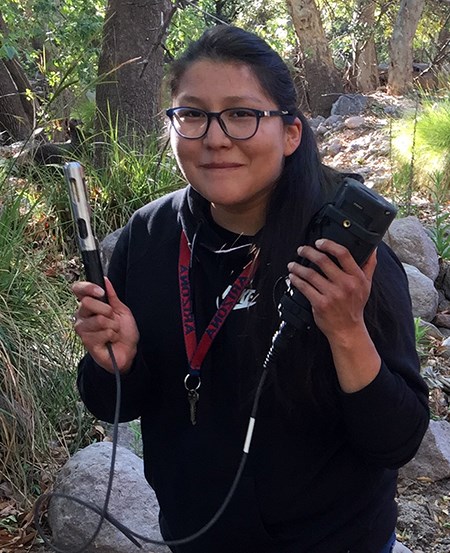 Black-haired woman wearing glasses stands holding water quality monitoring equipment