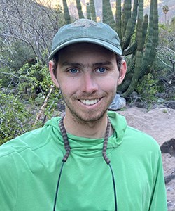 Smiling man with blue eyes and green shirt stands in front of cacti.