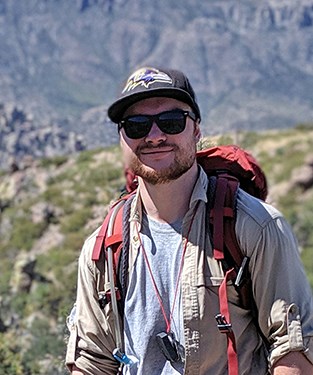 Bearded man in sunglasses stands on a desert mountainside