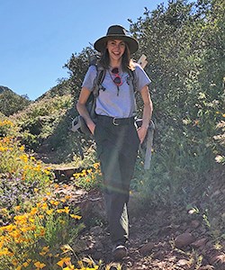 A woman in a hat walks on a trail surrounded by wildflowers
