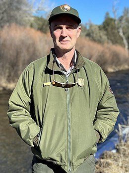 A National Park Service employee shown from the waste up in a uniform jacket and uniform cap near a river.