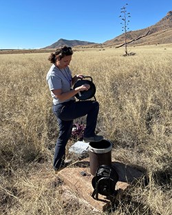 A scientist standing next to a metal, cylindrical well opening and  lowering a measuring tape into the well