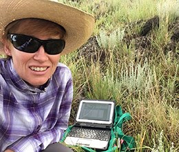 A scientist crouched down in a grassland next to a laptop computer.