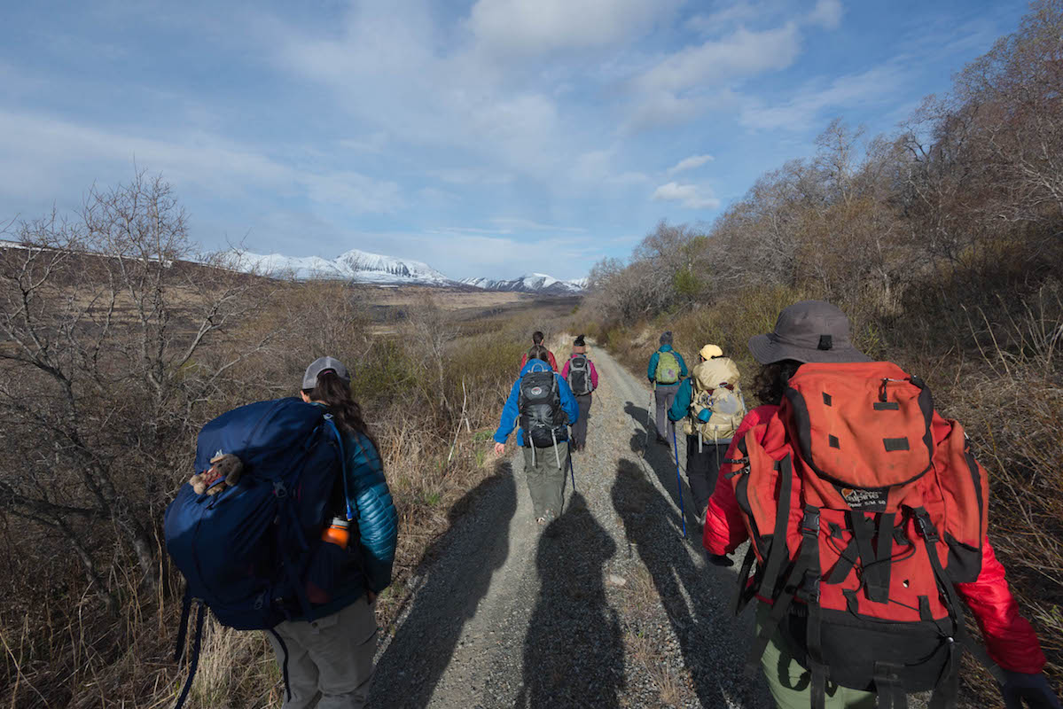 a group of people walk a dirt road towards snow covered mountains