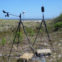 lt= A weather station attached to a tripod stands on a grassy sand dune next to a microphone on a tripod. Ocean and blue sky are in the background.