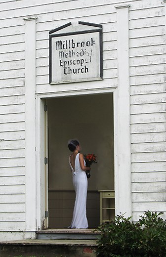 A bride in her white dress stands in the tall doorway of Millbrook Church, holding a bouquet of red roses upright against her front