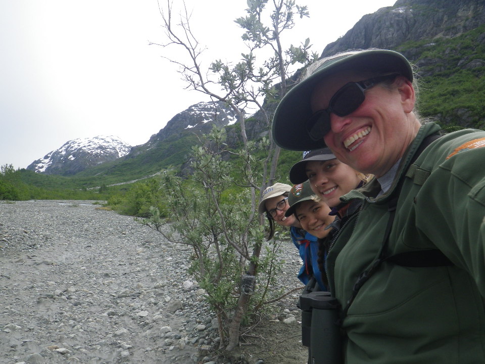 Laura Prugh (UW), Kiana Young (NPS), Mira Sytsma (UW) and Tania Lewis (NPS) pose by a recently installed motion sensor camera (on lower trunk of willow tree) in Tarr Inlet.