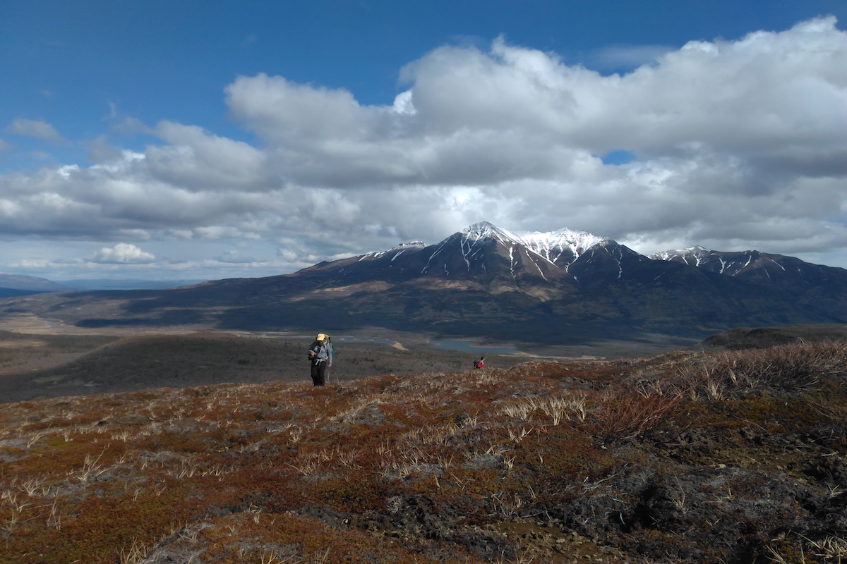 Two hikers climb a summit, with a mountain and lakes in the background