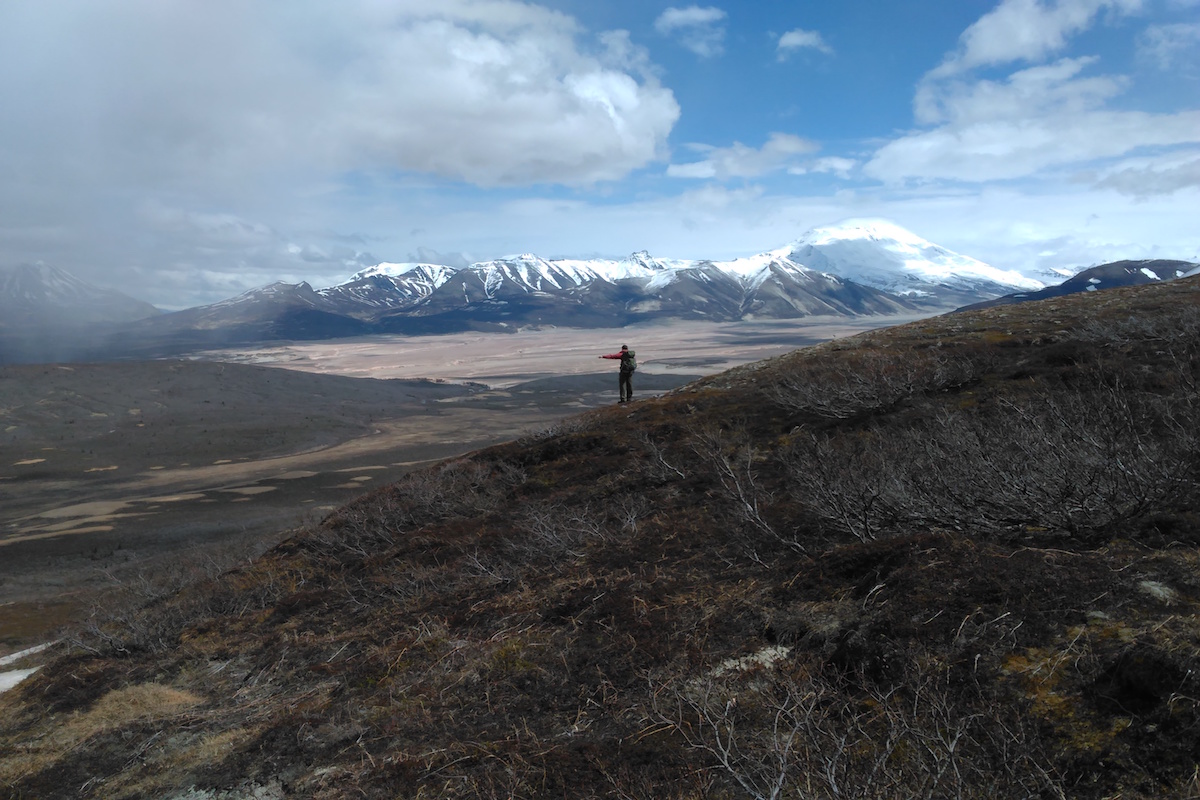 A hiker views a valley of ash deposits with rain clouds and mountains in the background.