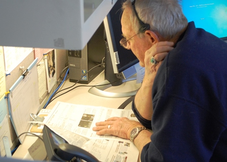Volunteer Jim Johnson consults the park newspaper to answer a visitor question. (NPS Photo / Will Sunderland) A volunteer looks at a newspaper and talks on the phone.