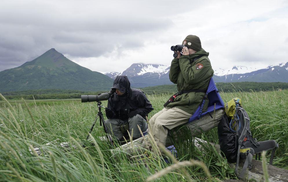 Two women look through binoculars while sitting in the grass