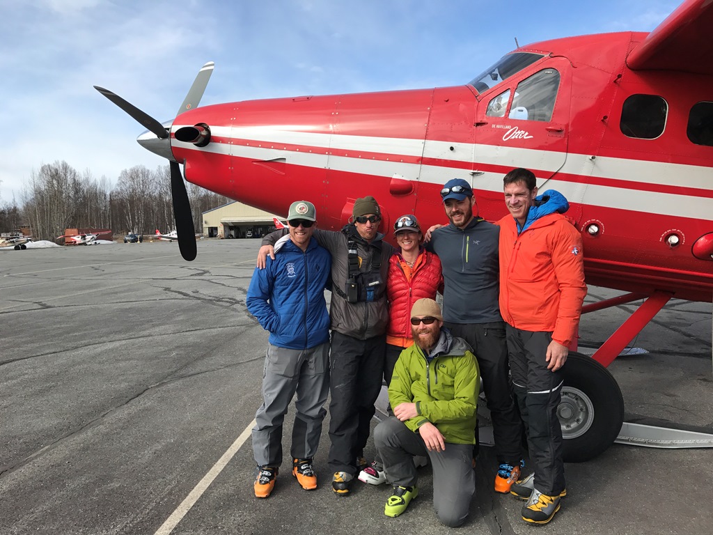A team of six climbers in mountain gear pose outside a small airplane