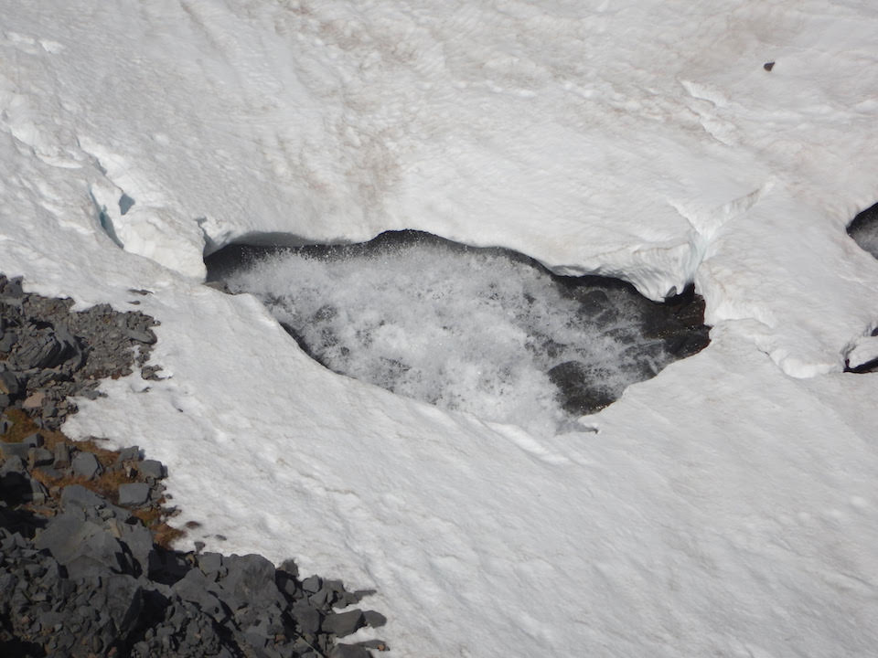 Close up view of a hole in snow showing rushing water underneath.