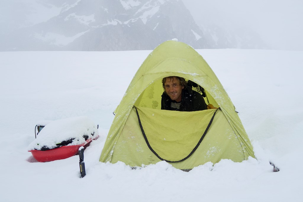 A smiling climber peaks out from a snowy tent