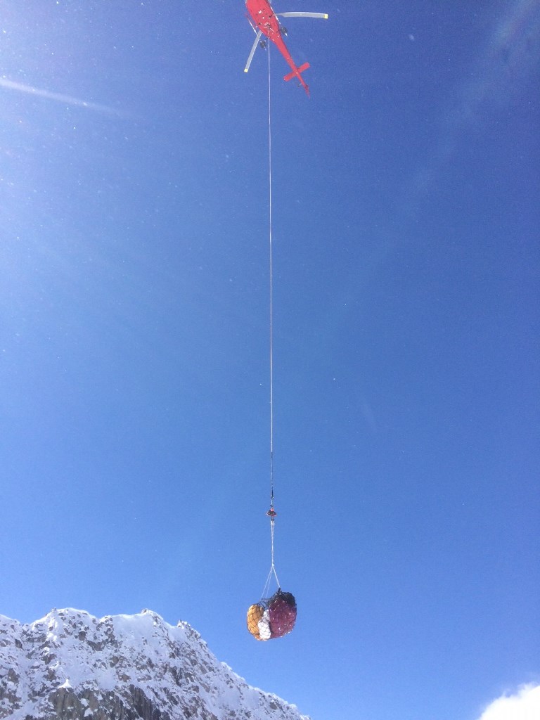 A helicopter lifts off basecamp with a slingload of camp gear